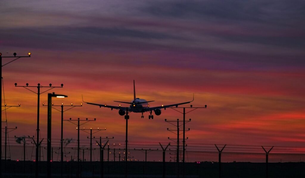 Airplane landing at sunset with vibrant red and purple skies, runway lights glowing, and silhouetted approach towers.
